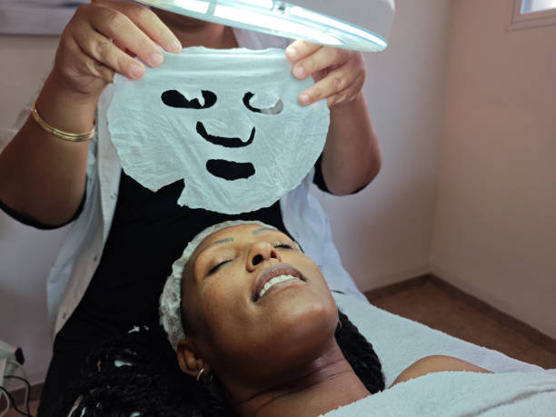 A spa therapist holds a delicate white sheet mask under a magnifying lamp, ready to apply it to the relaxed face of a client. The image captures a moment of rejuvenation and self-care, emphasizing professional skincare and hydration treatments.