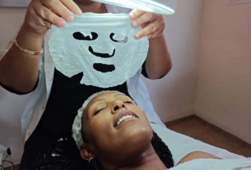 A spa therapist holds a delicate white sheet mask under a magnifying lamp, ready to apply it to the relaxed face of a client. The image captures a moment of rejuvenation and self-care, emphasizing professional skincare and hydration treatments.